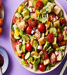 A colorful salad with cherry tomatoes, cucumber slices, lettuce, yellow bell pepper, radish cubes, croutons, and green onions, served on a white plate against a purple background. GREENHOUSE Goodness