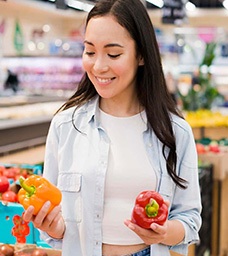 A woman with long dark hair smiles while holding a yellow bell pepper in one hand and a red bell pepper in the other at a grocery store produce section. GREENHOUSE Goodness